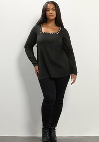 Maglia nera a maniche lunghe con scollo quadrato, decorata con borchie circolari. Abbinata a pantaloni neri aderenti e stivaletti. Texture liscia.