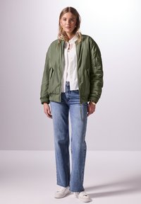 Olivgrön bomberjacka med en slät yta, utrustad med en dragkedjeficka på ärmen, i kombination med ljusblå raka jeans och vita sneakers.