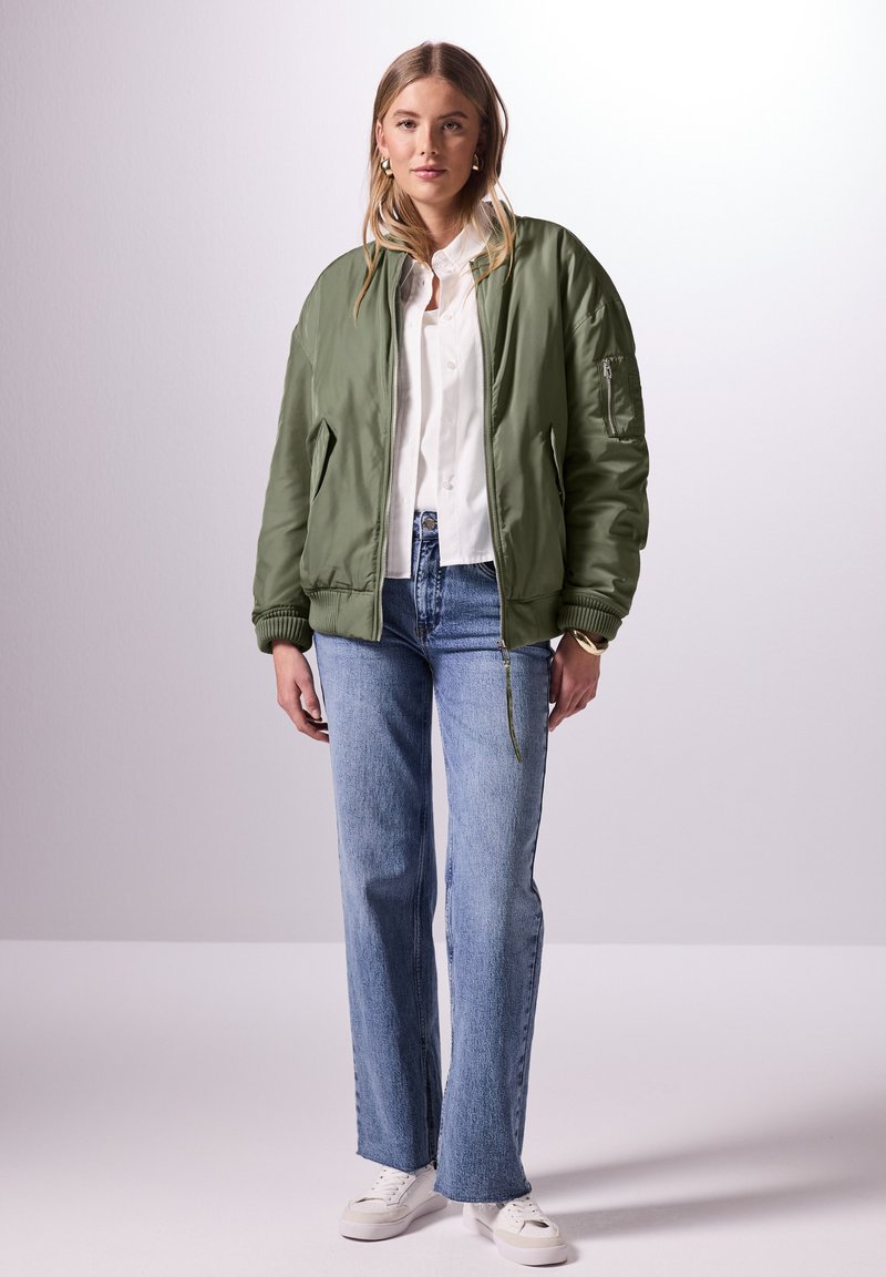 Olivgrön bomberjacka med en slät yta, utrustad med en dragkedjeficka på ärmen, i kombination med ljusblå raka jeans och vita sneakers.