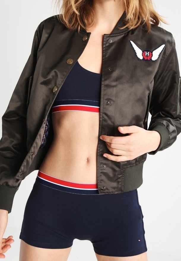 Femme portant une veste bomber sombre ouverte avec un emblème ailé sur un soutien-gorge de sport bleu marine et un short assorti avec des rayures rouges et blanches.