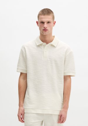 Jonge man draagt een gestructureerd crèmekleurig poloshirt met korte mouwen en bijpassende korte broek, staand tegen een effen witte achtergrond.