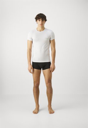 Camiseta blanca ajustada con un pequeño logotipo verde, combinada con calzoncillos boxers negros. El modelo está descalzo frente a un fondo sencillo.