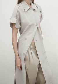 Jasno beżowy trencz z lnu o podwójnym zapięciu, z podwiniętymi rękawami i dużymi guzikami, założony na dopasowane spodnie.