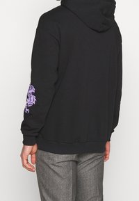Persona con un hoodie nero decorato con dragoni viola sulle maniche e pantaloni a scacchi grigi, in piedi contro uno sfondo bianco semplice.