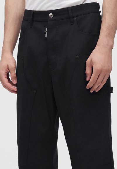 Gros plan d'une personne portant un pantalon cargo noir avec bouton et fermeture éclair, plusieurs poches, et une chemise blanche rentrée.