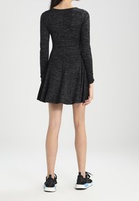 Donna in piedi, che indossa un vestito knit a maniche lunghe di colore grigio scuro con una gonna svasata e sneakers nere con suole bianche e blu.