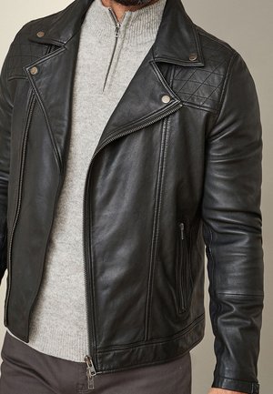 Homme portant une veste en cuir noire par-dessus un pull gris clair zippé et un pantalon foncé, visible des épaules jusqu'aux hanches.