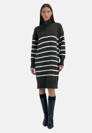 Kvinde iført en knælængde sort sweaterkjole med hvide vandrette striber, en halv-zip krave og sorte knæhøje læderstøvler.