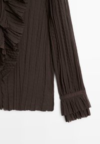Blouse brune plissée avec des détails froncés sur le devant. Manches longues avec des poignets boutonnés et un tissu texturé. Matériau doux et léger.