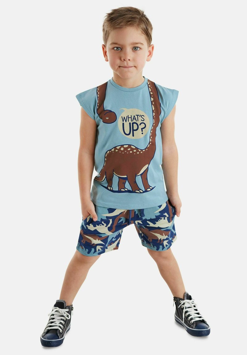 Blaues ärmelloses T-Shirt mit einem Dinosaurier-Grafik und dem Text "WAS GIBT'S?", kombiniert mit gemusterten blau-braunen Camouflage-Shorts; schwarze High-Top-Sneaker.
