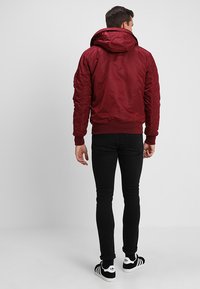 Homemem vestido com uma jaqueta bomber com capuz em cor bordeaux, jeans skinny pretos e ténis Adidas em preto e branco, de costas contra um fundo simples.