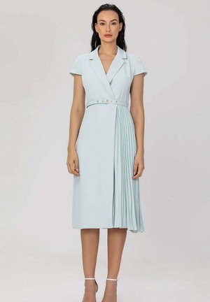 Femme portant une robe ceinturée bleu clair à manches courtes, col à revers, avec un détail plissé sur un côté, debout devant un fond uni.