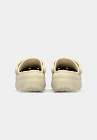 Sabots beiges au design lisse et arrondi, dotés de trous de ventilation et de brides arrière. La semelle présente des détails texturés pour une meilleure adhérence.