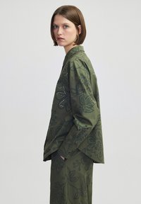 Olijfgroene blouse met bloemenborduursel en textuuraccenten. Losse pasvorm, lange mouwen en een puntkraag. Gecombineerd met bijpassende broek.