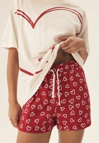 Czerwone szorty w białe wzory serduszek, z wiszącym paskiem w talii, połączone z luźną kremową koszulką z czerwonymi zakrzywionymi liniami. Materiał z mieszanki bawełny.