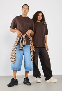 Dos personas llevan camisetas marrones de gran tamaño. Una de ellas combina una camisa de cuadros alrededor de la cintura. Ambas las emparejan con pantalones cortos de mezclilla y pantalones de pierna ancha.