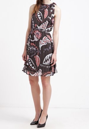 Vrouw die een mouwloze, geplooide jurk draagt met een zwart-wit-roze bloemmotief, gecombineerd met zwarte pumps met een spitse neus.