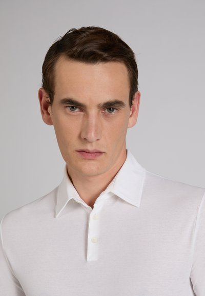 Jeune homme aux cheveux bruns courts portant une chemise blanche à col, regardant droit devant avec une expression neutre sur un fond gris uni.