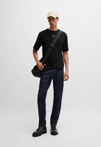 Czarna koszulka z okrągłym dekoltem z logo "BOSS", zestawiona z ciemnoniebieskimi dżinsami. Nosi beżową czapkę i niesie czarną torbę na ramię. Czarne buty.