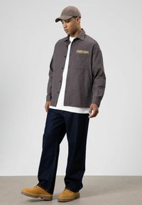 Bruin en zwart geruit overhemd met een zak en de tekst "FREEDOM", gedragen over een wit shirt en gecombineerd met donkere spijkerbroek en beige schoeisel.