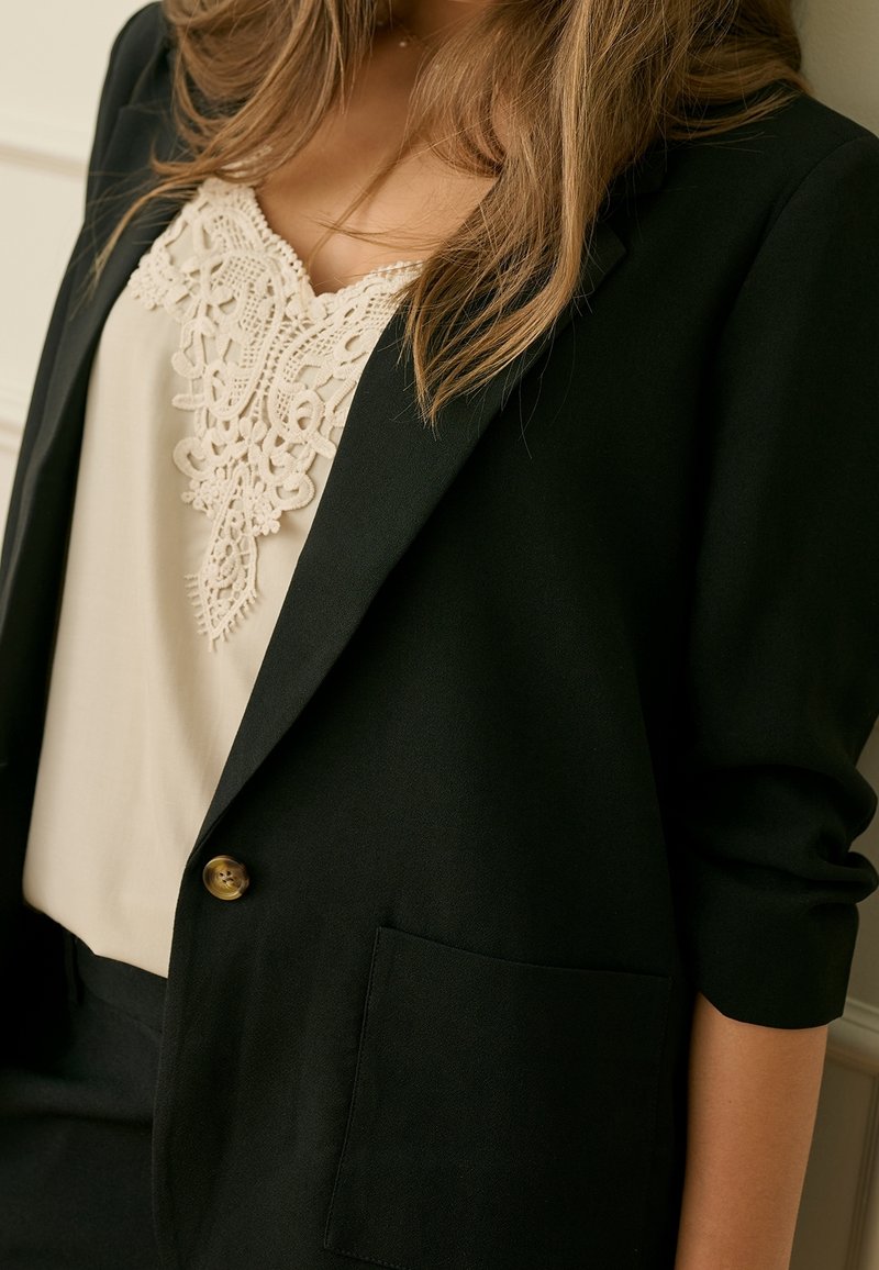 Femme portant un blazer noir sur un chemisier crème avec un détail en dentelle complexe, manche du blazer légèrement relevée, cheveux bruns tombant sur les épaules.