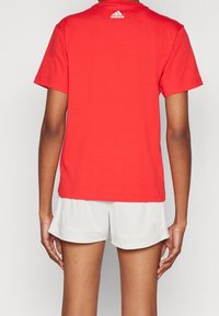 Röd bomullströja med korta ärmar, med en vit Adidas-logga nära halsen, kombinerad med ljusgrå shorts.