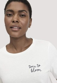 Witte katoenen t-shirt met ronde hals, featuring marineblauwe cursieve borduurwerk met de tekst "tijd om te bloeien" aan de linkerkant.