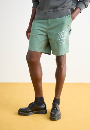 Grüne Baumwollshorts mit einem grafischen weißen Gesichtdesign, vorderen Taschen und einem entspannten Schnitt. Kombiniert mit schwarzen Schuhen und Socken.