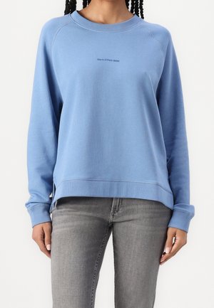 Personne portant un sweat-shirt bleu clair à col rond avec un petit texte sur la poitrine et un jean gris, debout devant un fond blanc uni.