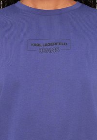 Fialové bavlněné tričko s kulatým výstřihem a černým potiskem loga, které nápis "KARL LAGERFELD JEANS" přetíná hrudník.
