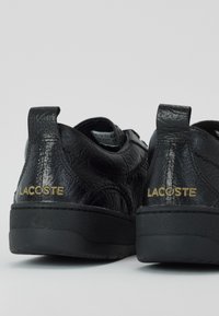 Juodi odiniai batai su tekstūruotu paviršiumi, turintys žemą apykaklę, juodus guminis padas ir auksinę "Lacoste" ženklinimą ant kulno.