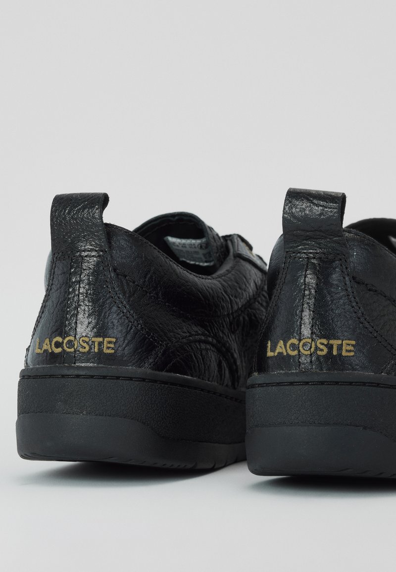 Juodi odiniai batai su tekstūruotu paviršiumi, turintys žemą apykaklę, juodus guminis padas ir auksinę "Lacoste" ženklinimą ant kulno.