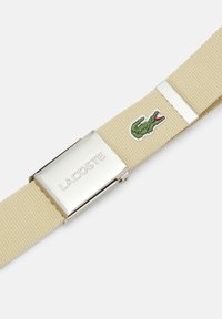 Béžový tkaný opasok so striebornou kovovou prackou s gravírovaním "LACOSTE" a nášivkou zeleného krokodíla. Odolný materiál a elegantný dizajn.