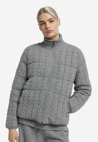 Veste grise en maille torsadée avec un col montant, une fermeture éclair frontale et deux poches. Présente des motifs texturés ainsi que des poignets et un ourlet côtelés.