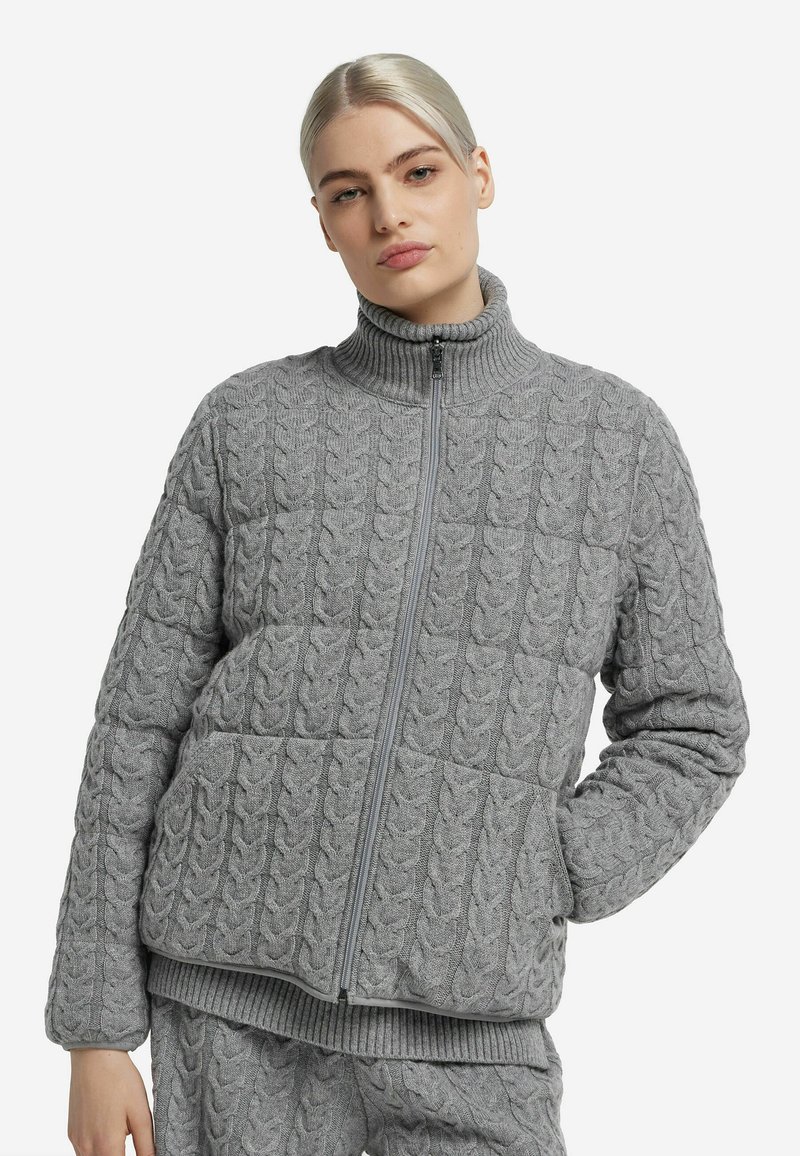 Veste grise en maille torsadée avec un col montant, une fermeture éclair frontale et deux poches. Présente des motifs texturés ainsi que des poignets et un ourlet côtelés.