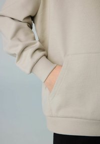 Sudadera beige con puños de canalé y un bolsillo tipo canguro, exhibida sobre un fondo claro con textura visible.
