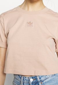 Lyhythihainen lyhyt t-paita vaaleanpunaisesta puuvillasta, jossa on ommeltu logo rinnassa ja teksturoidut raidoitusyksityiskohdat olkapäissä.