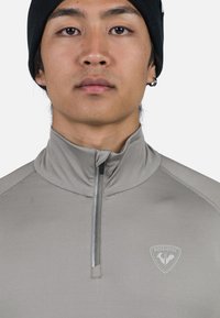 Ung person som bär en svart beanie och en grå zip-up-tröja med Rossignol-logotyp, vänd framåt mot en enfärgad bakgrund.