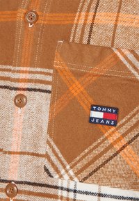 Brązowa flanelowa tkanina w kratę z pomarańczowymi i czarnymi paskami. Zawiera kieszeń na piersi z naszywką logo "Tommy Jeans" oraz brązowe guziki.