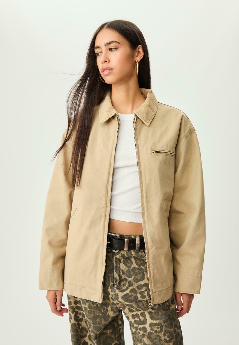 Ung kvinde med langt mørkt hår, iført en beige jakke, hvid crop top, leopardmønstrede bukser og sort bælte, der kigger til siden mod en lys baggrund.