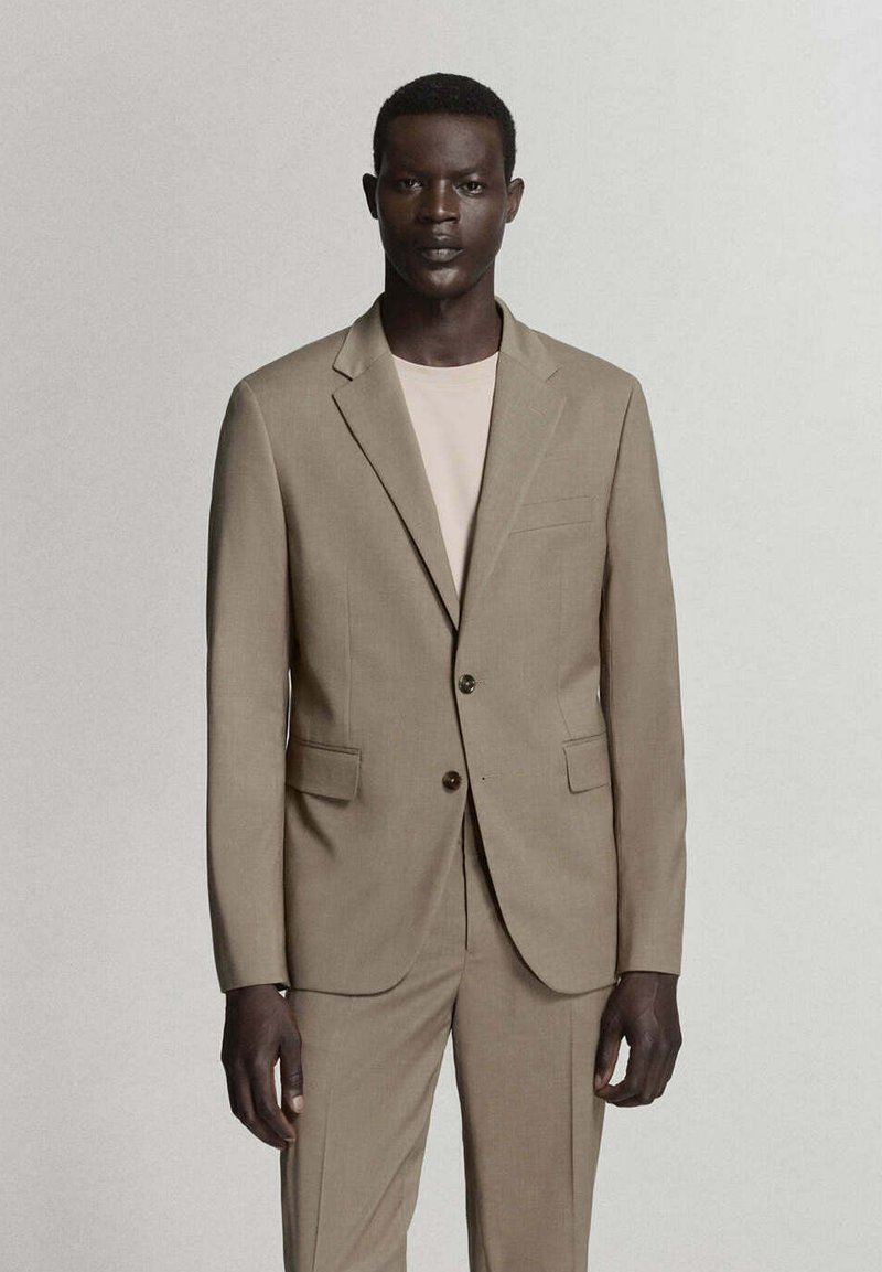 Homme à la peau foncée portant une veste de costume beige et un pantalon assorti sur une chemise beige clair, debout devant un fond uni.