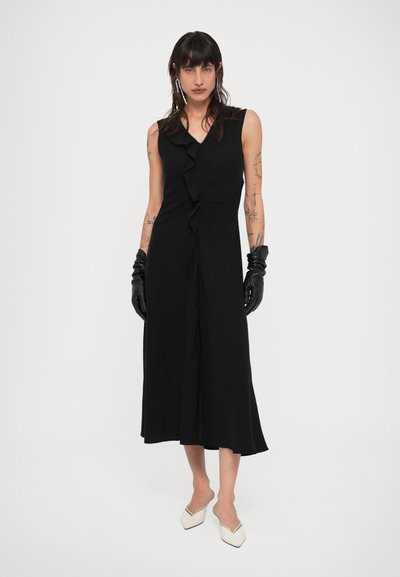 Sieviete ar gariem tumšiem matiem, tērpusies melnā piedurknēm neapsegta midi kleitā, melnās ādas cimdiņos, baltos augstpapēžu apavos ar smailiem galiņiem, redzamas tetovējumu zīmes uz rokām.