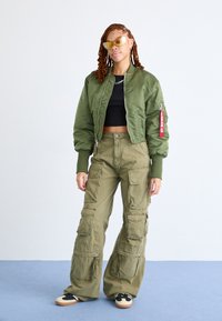 Olijfgroene bomberjack met geribde manchetten, zwarte crop top, olijfgroene cargobroek met meerdere zakken en zwarte sneakers met lichte zolen.