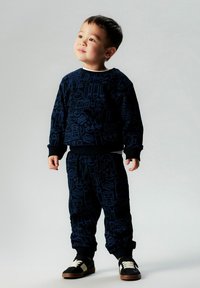 Marineblå joggedress for barn med mønsterdesign, bestående av en genser med rund hals og bukser med elastisk midje. Kombinert med svarte joggesko.