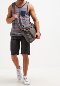 Sac messager gris avec des sangles marron, porté en bandoulière. Modèle portant un débardeur rayé et un short, avec des baskets blanches. Cadre décontracté en extérieur.