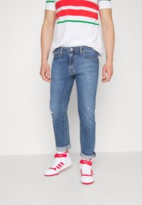 Modré džínové slim fit kalhoty s efektem opotřebení. Kombinovány s bílým pruhovaným tričkem a červeno-bílými teniskami.