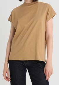 Camiseta corta de manga larga color marrón, hecha de algodón suave, con un corte relajado y un escote alto, llevada con jeans oscuros. Sin patrones ni acentos visibles.