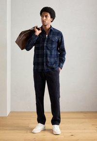 Man in een marineblauw geruit shirt, donkere broek, witte sneakers, met een bruin leren duffel bag over zijn schouder, staande op een houten vloer.