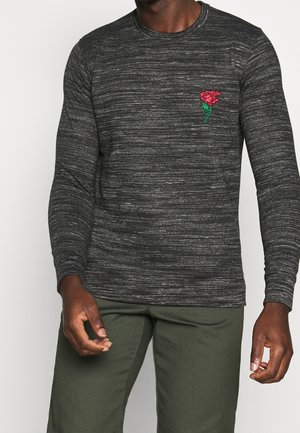 Mann trägt ein schwarzes und graues gestreiftes Langarmshirt mit einer roten gestickten Rose auf der Brust und grüne Hosen.