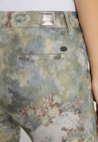 Pantalons en denim à motifs floraux dans des teintes vert clair, bleu et crème. Comprend une poche arrière, un patch en cuir et un tissu texturé.